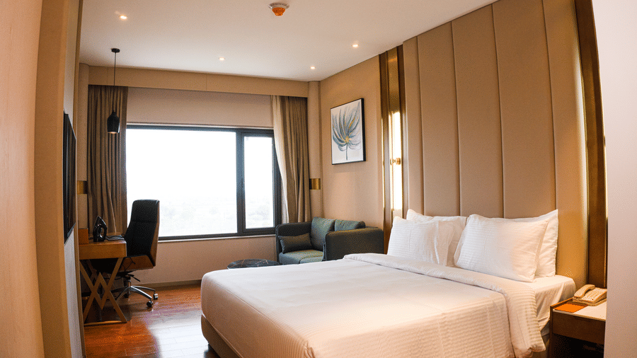 A king size bed, cushioned sofa chair with a coffee table, and an office desk unit under a window at Pride Plaza Indore.