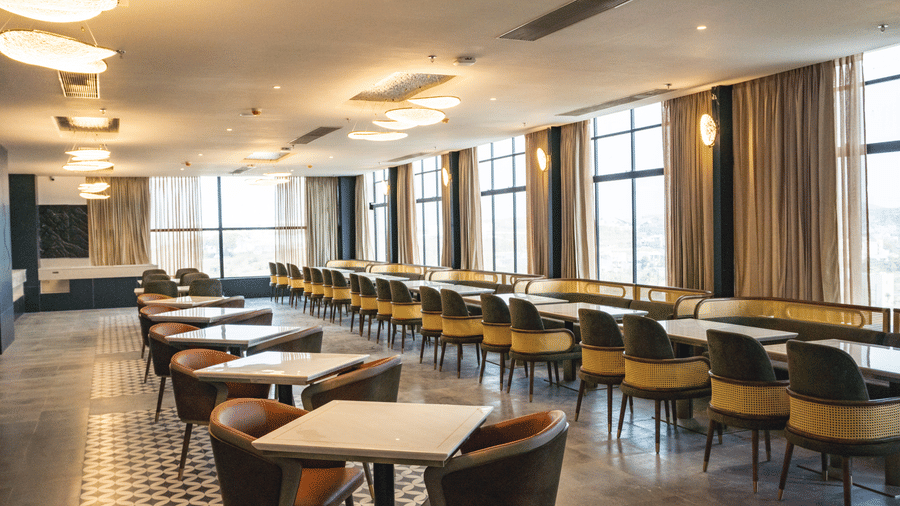 A spacious dining hall with square tables, upholstered chairs, and ceiling lights at Pride Plaza Indore.