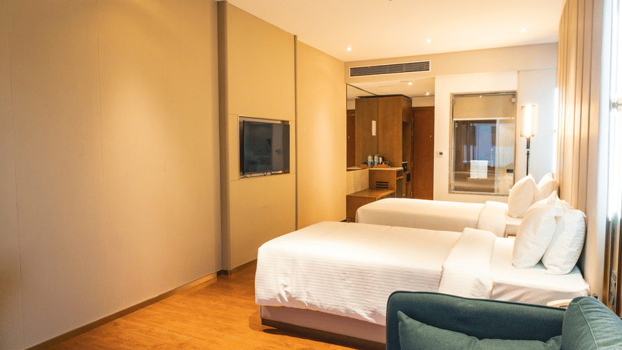 Single beds, a television screen mounted on the wall, and a corner couch on a polished wooden floor at Pride Plaza Indore.