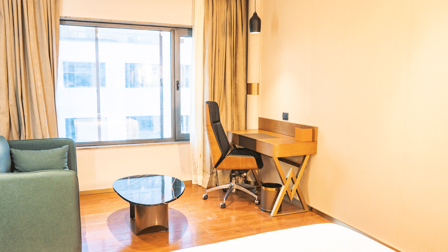 A glass coffee table, sofa chair, and wooden desk with a wheeled office chair positioned next to a window at Pride Plaza Indore.