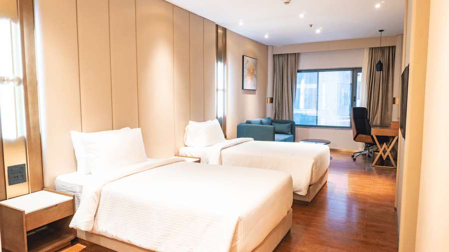 Single beds made with linens, a corner sofa chair, and a study desk overlooking a window at Pride Plaza Indore.