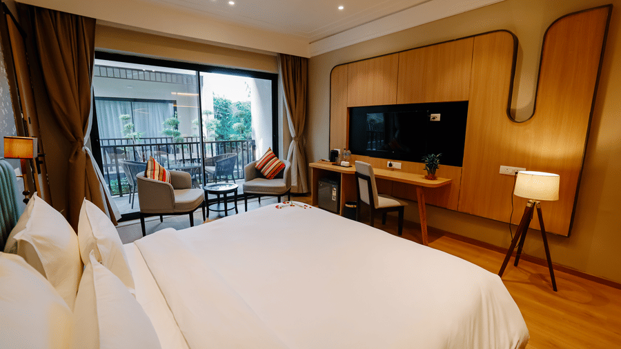 Pride Premier Alwar | A room with a large bed, bedspreads, pillows, a television, windows showcasing a private balcony