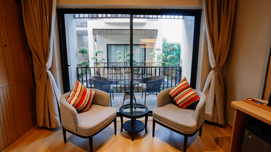 Pride Premier Alwar | A cottage living room with pillows on chairs, and windows covered with curtains in the backdrop.