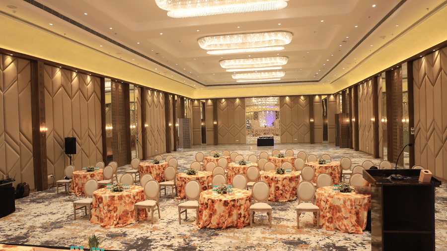 Pride Premier Alwar- Spacious, brightly lit grand ballroom set up with numerous circular banquet tables and ornate carpeting.