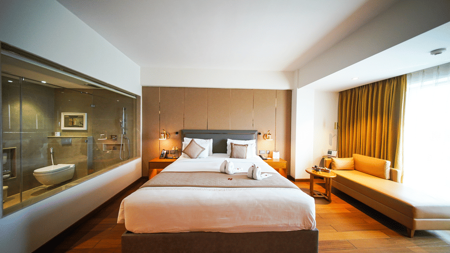 An interior view of the Premium King Room at Pride Premier Udaipur. A double bed sits in the centre with a daybed placed by the window curtains, whilst a glass wall on the left looks into the bathroom area.