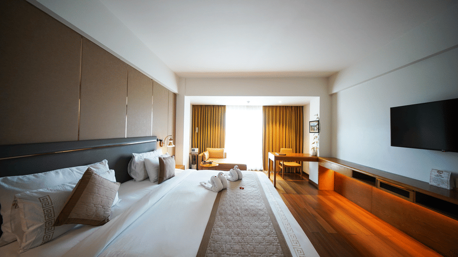A view across the Premium King Room at Pride Premier Udaipur from the side of the bed. The image shows a double bed with linen and pillows, a wall-mounted television, a desk, and a seating area positioned near the window at the far end.