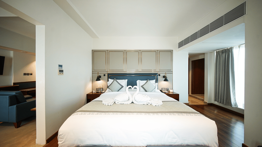 An interior view of a bedroom section within the Premium Suite Room at Pride Premier Udaipur. A double bed with pillows and a folded fabric arrangement sits in the centre of the room, positioned between bedside units.
