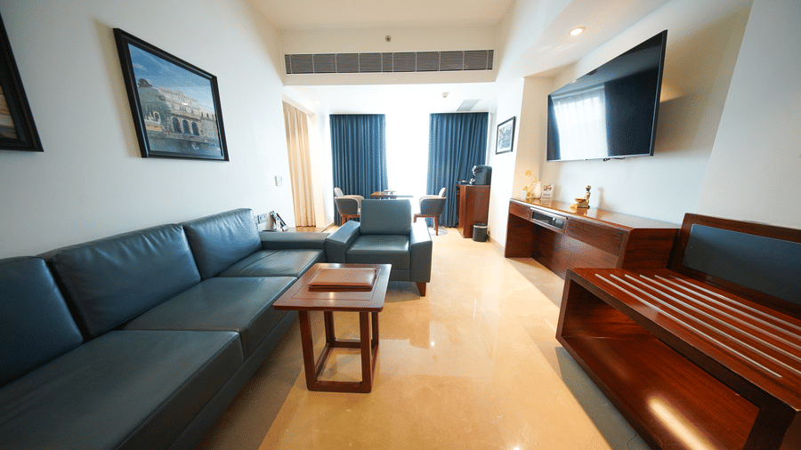 A view across the lounge area of the Premium Suite Room at Pride Premier Udaipur. The space is furnished with a sofa, a coffee table, a television mounted on the wall, and cabinetry along the side.