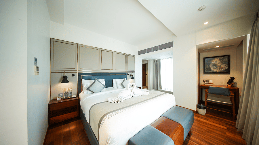 An angled view of the bedroom area in the Premium Suite Room at Pride Premier Udaipur. The space displays a double bed with linen, bedside tables, lamps, and an opening leading towards a desk and chair.