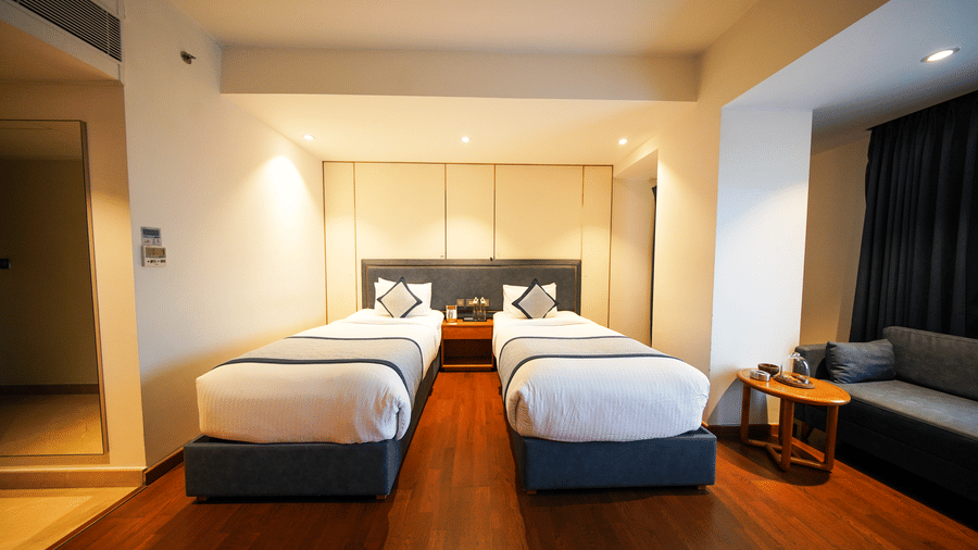 A front-facing view of the bedroom area in the Premium Twin Room at Pride Premier Udaipur. Two single beds are placed side by side with a table between them. A sofa sits on the right side next to window drapes, and the floor is made of timber.
