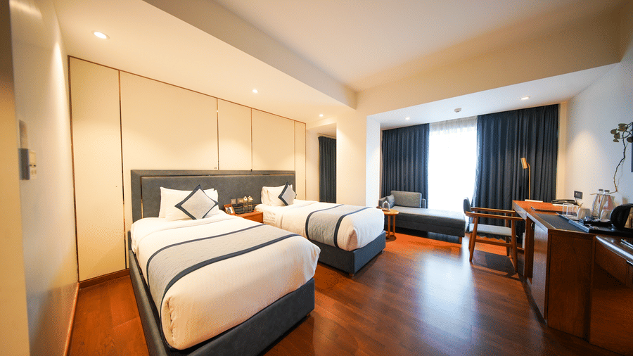An angled view across the Premium Twin Room at Pride Premier Udaipur. The space displays two single beds arranged in sequence along the wall. A desk with a chair and a wall fixture are positioned on the opposite side near the window area.
