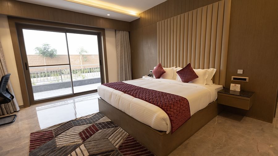 A stylish hotel room at Pride Resort, Jodhpur, featuring a large bed with cushions and a balcony with a view outside.