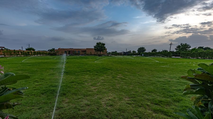 A lush green lawn with sprinklers watering the grass at Pride Resort, Jodhpur, under a cloudy evening sky.