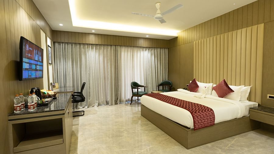 A well-lit hotel room at Pride Resort, Jodhpur, with a large bed, TV, desk, and seating area by the window.