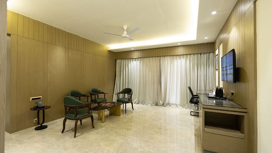 A spacious seating area with chairs and a desk opposite a wall-mounted TV at Pride Resort, Jodhpur, with large windows.