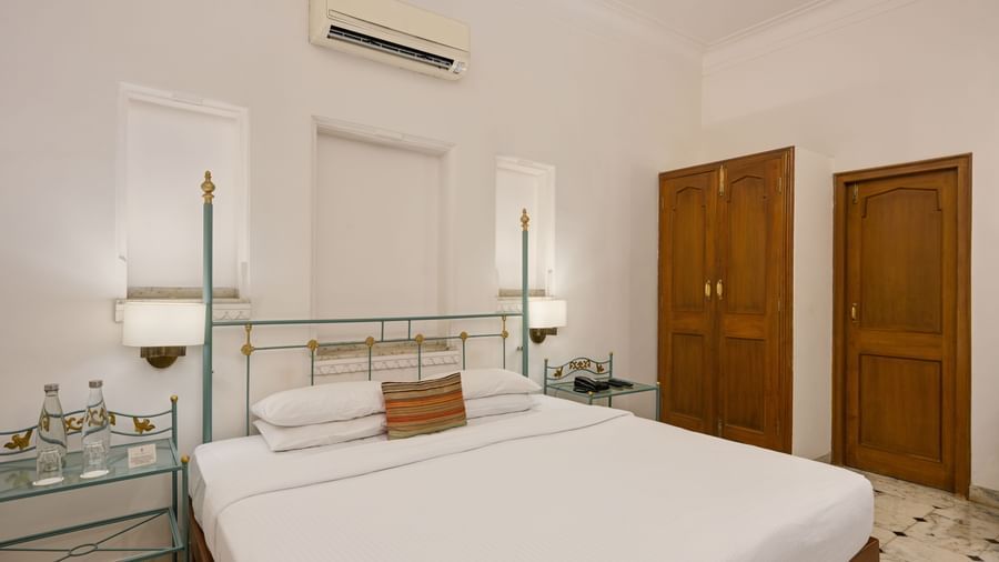 A bedroom at Ram Pratap Palace in Udaipur, featuring a well-made bed, glass-topped side tables, a wooden wardrobe, and an air-conditioning unit mounted above the bed.