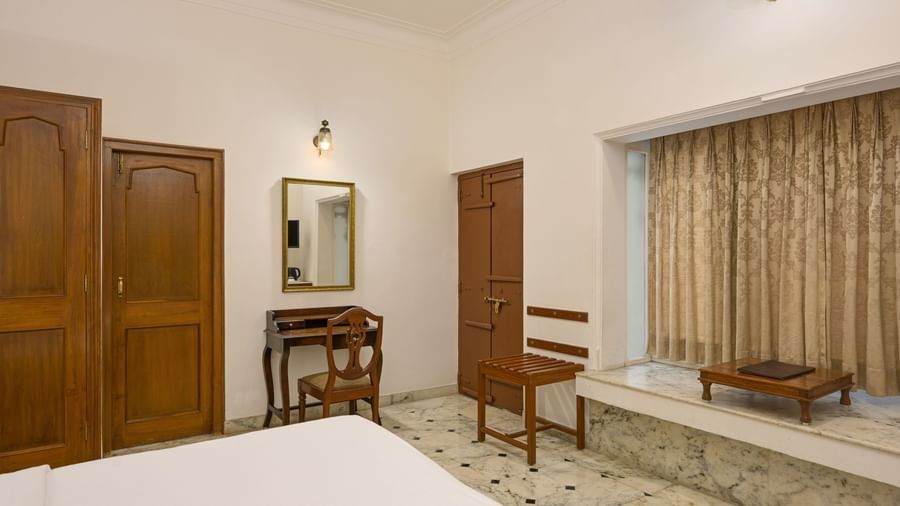 A room at Ram Pratap Palace in Udaipur, furnished with a bed, a wardrobe, a desk with a chair, a mirror, a small stool, a window seat with curtains, and a low wooden table set on a marble platform.