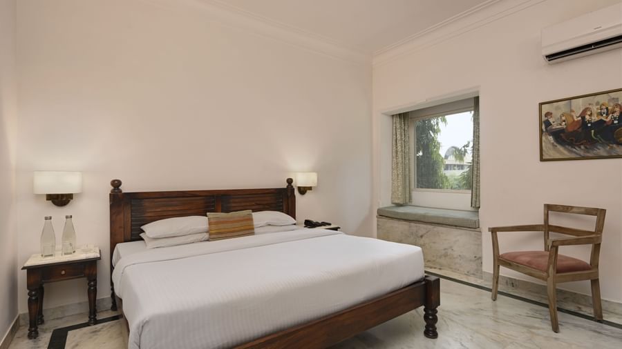 A well-lit room at Ram Pratap Palace, Udaipur, with a large wooden bed, two side tables with lamps, a chair, and a window seat overlooking the property.