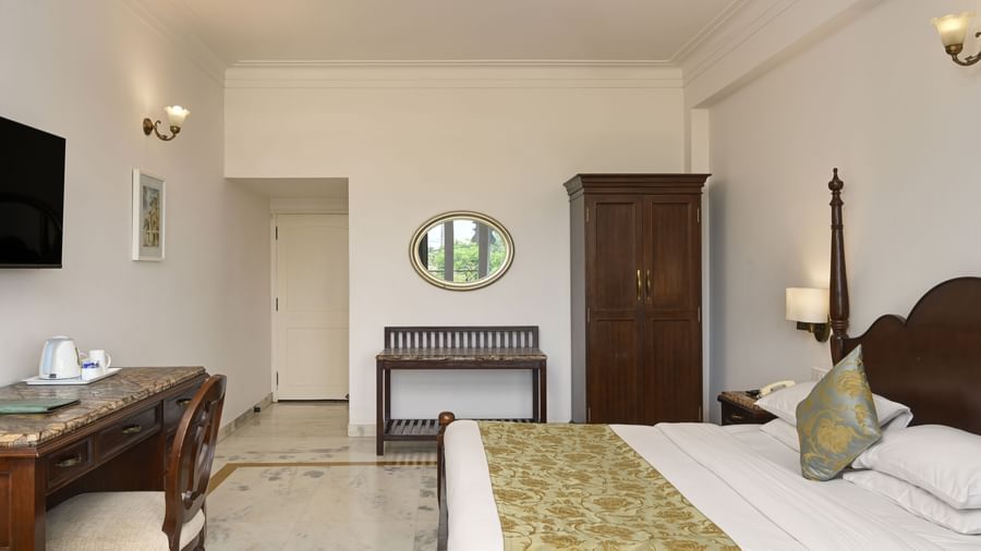 A room at Ram Pratap Palace, Udaipur with a decorated wooden bed, bedside table, wall-mounted TV, tea/coffee station with chair, wardrobe, and vanity.