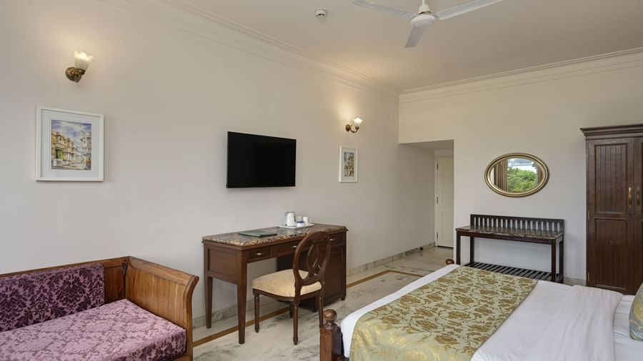 A hotel room at Ram Pratap Palace with a bed, a daybed, a desk, a TV, a mirror, and a wardrobe, all on a light-coloured tile floor.