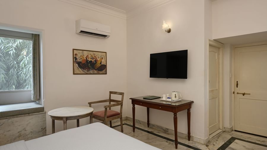 A room at Ram Pratap Palace in Udaipur, furnished with a bed, a television, air-conditioning, a wall painting, a wooden desk, a small round table with a chair, and a window seat overlooking lush greenery.