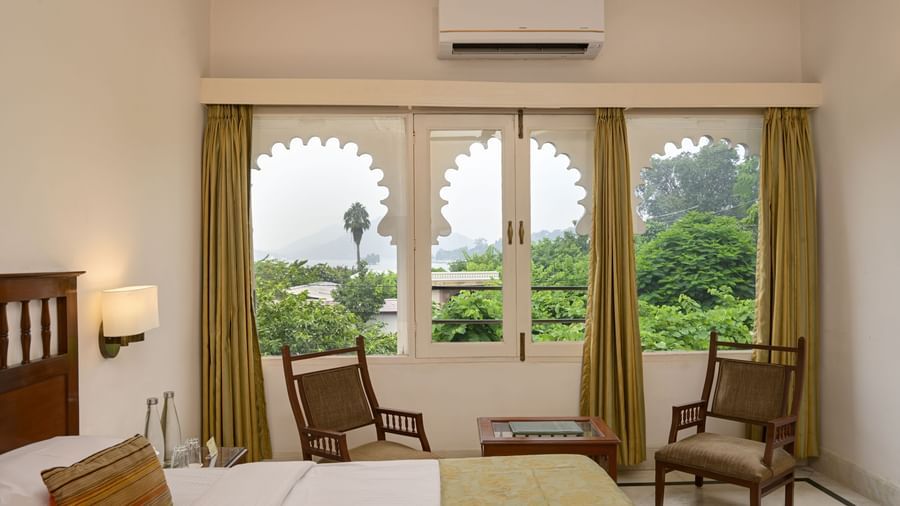 A guest room at Ram Pratap Palace in Udaipur, with a bed, two chairs, a side table, and a window with arched frames overlooking the outdoors.