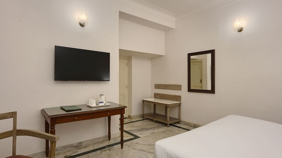 A minimalist room at Ram Pratap Palace in Udaipur, featuring a bed, a wall-mounted TV, a wooden desk with a tea and coffee maker and a menu, a bench, a mirror, and marble flooring.