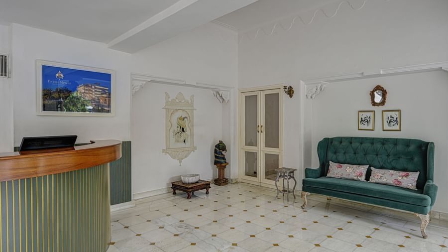 Reception area of Ram Pratap Palace, Udaipur with a curved wooden desk, green sofa with cushions, wall paintings, decorative statue, and double door.