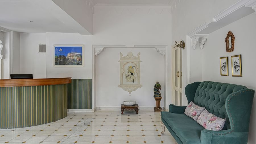 Reception area of Ram Pratap Palace, Udaipur with a wooden desk, green upholstered sofa with cushions, wall art, framed pictures, and decorative statue.