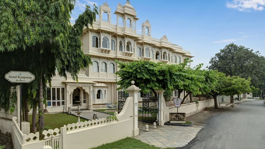 Exterior view of Ram Pratap Palace, Udaipur showing a multi-storey heritage-style building with arched windows, entrance gate, garden area, and surrounding trees.