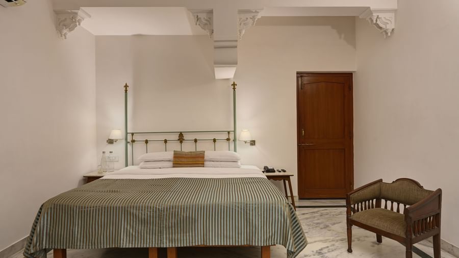 A bedroom at Ram Pratap Palace in Udaipur, featuring a four-poster bed with a striped bedspread, a small chair, bedside tables, and two wall-mounted lamps flanking the bed.