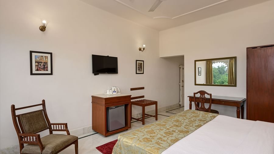 A hotel room at Ram Pratap Palace in Udaipur, with a bed, a chair, a mini fridge, a wall-mounted TV, a desk, and a large mirror.