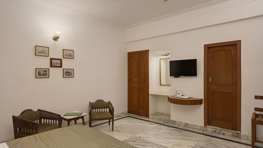 A room at Ram Pratap Palace in Udaipur, featuring a bed, a wall-mounted TV, two wooden chairs with a table, a tea and coffee maker placed on a wooden floating countertop, and a vanity area with a mirror.