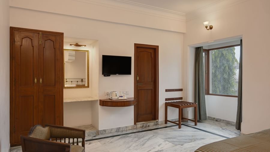 A bedroom at Ram Pratap Palace in Udaipur, featuring a large wooden wardrobe, a TV, a luggage rack, a chair, a vanity with a mirror, and a window with curtains overlooking greenery.