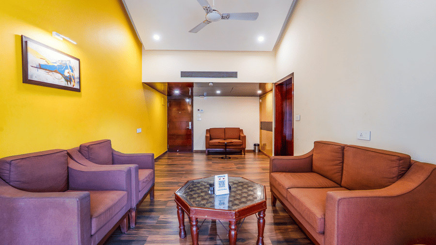 Bright living room of Rosetum Anjuna's Royal Suite with yellow accent wall, abstract artwork, multiple sofas, glass-top coffee table, and dark hardwood floors.