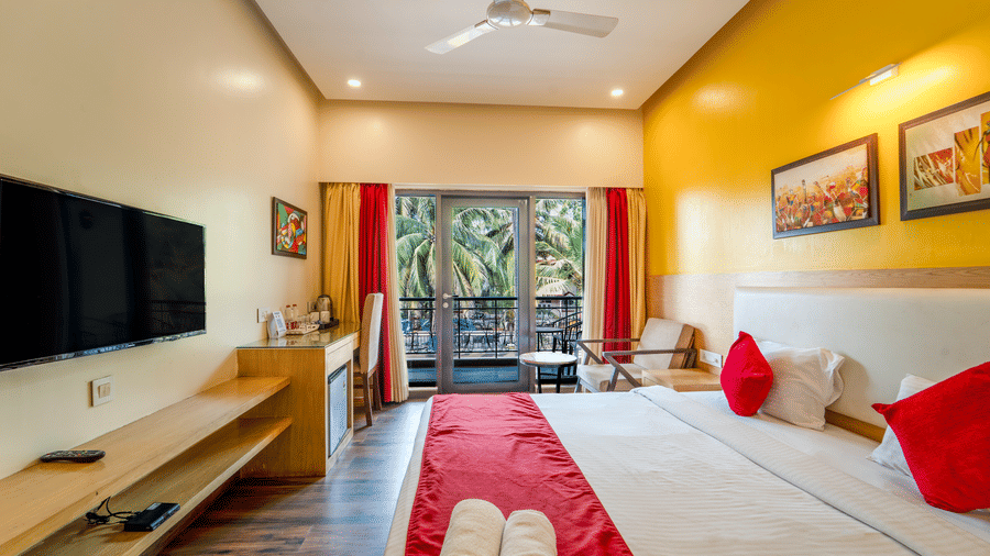 Rosetum Anjuna Deluxe Room with a king bed, red runner, rolled towels, yellow accent wall, wall-mounted TV, work desk with kettle, and balcony with palm tree views.