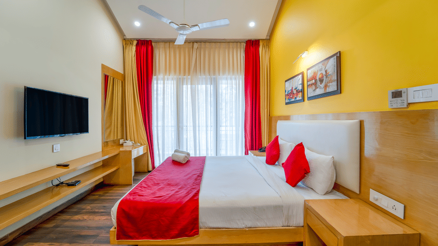 Rosetum Anjuna Deluxe Room with a king bed, red runner and pillows, yellow accent wall, framed artwork, flat-screen TV, sheer curtains, and a wooden bedside unit.