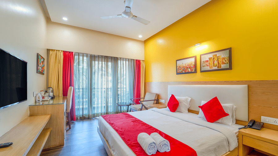 Full view of Rosetum Anjuna's Deluxe Room with king bed, rolled towels, red pillows, yellow accent wall, flat-screen TV, tea station, balcony seating, and framed artwork.