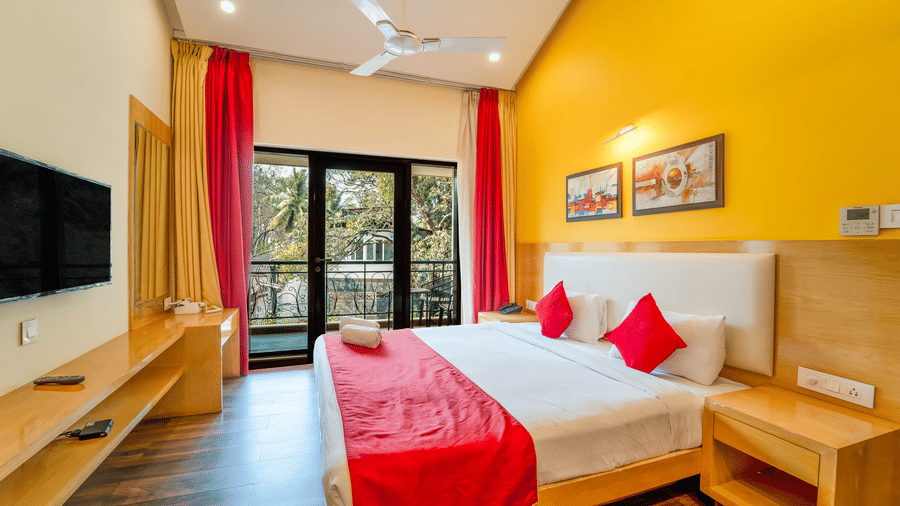 Rosetum Anjuna Deluxe Room with a king bed, red runner, red accent pillows, yellow walls, framed artwork, flat-screen TV, and open balcony doors with garden views.