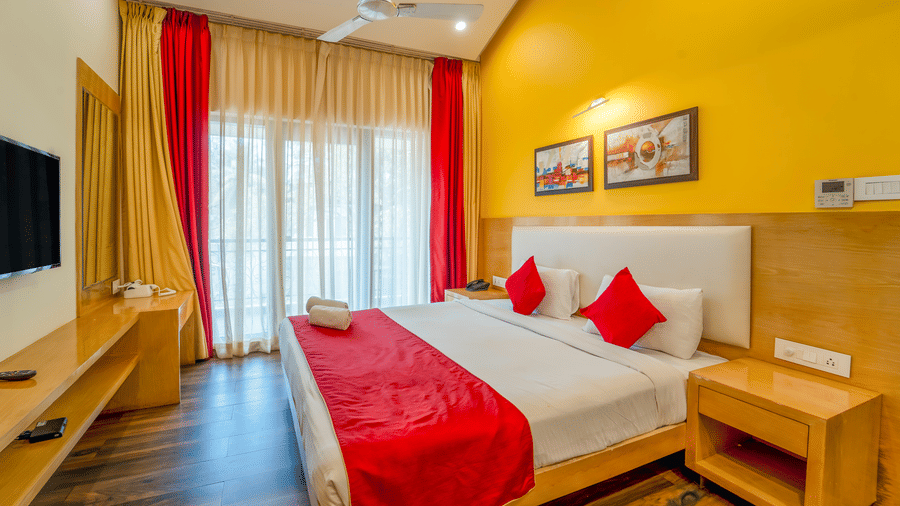 Rosetum Anjuna Deluxe Room featuring a king bed with red pillows and rolled towel, yellow feature wall, framed art, flat-screen TV, and red and gold floor-length curtains.
