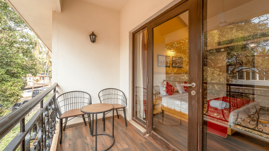 Private balcony of a Rosetum Anjuna Deluxe Room with two iron chairs, a round table, wrought iron railings, and a garden view with palm trees, overlooking the room interior.