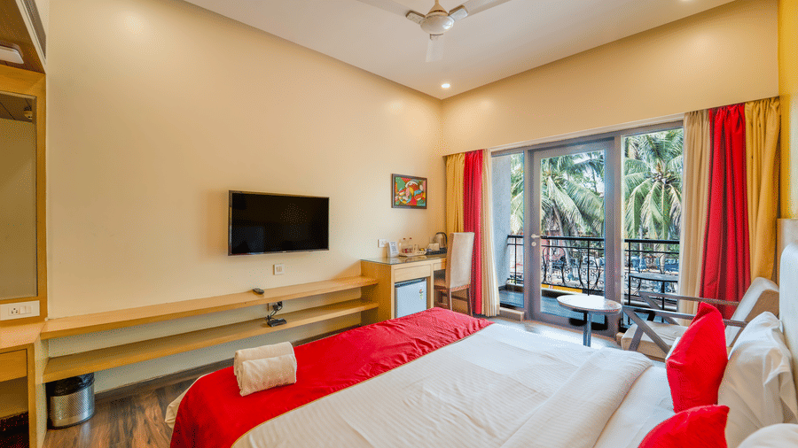 Rosetum Anjuna Deluxe Room showing king bed with red pillows, flat-screen TV on wooden console, balcony with outdoor seating, red and gold curtains, and palm tree views.