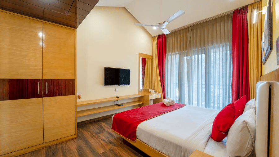 Rosetum Anjuna Deluxe Room with built-in wooden wardrobe, king bed with red accent, wall-mounted TV, dressing mirror, sheer curtains, and large window with natural light.