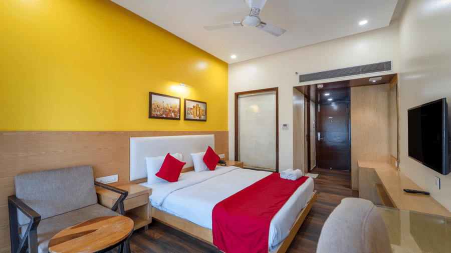Rosetum Anjuna Deluxe Room with a king bed, red accent runner, yellow feature wall, framed artwork, flat-screen TV, lounge chair, and glass-panelled bathroom entrance.