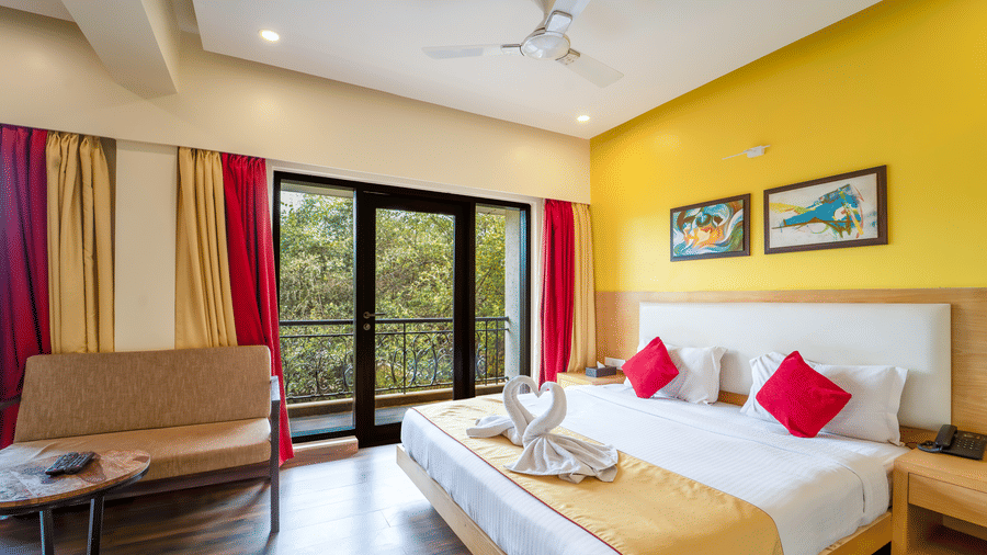 Rosetum Anjuna's Executive Suite bedroom featuring a king bed with towel swans, red cushions, balcony with garden view, yellow accent wall, and framed artwork above the headboard.