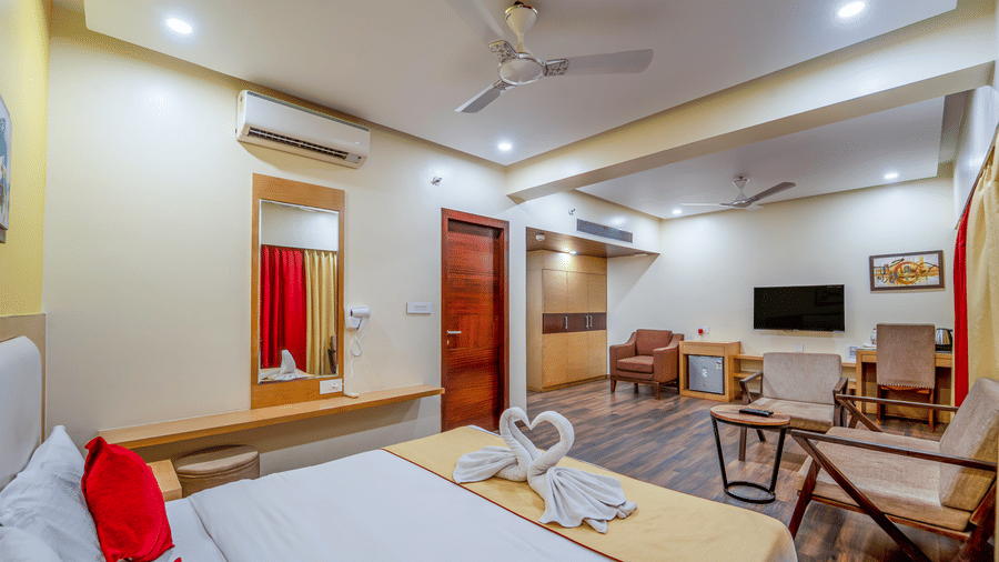 Wide-angle view of Rosetum Anjuna's Executive Suite with king bed, towel swans, red pillows, wall mirror, built-in wardrobe, seating area, and wall-mounted TV.