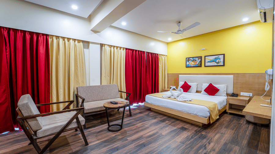 Wide-angle view of Rosetum Anjuna's Executive Suite with king bed, towel swans, red pillows, wall mirror, built-in wardrobe, seating area, and wall-mounted TV.