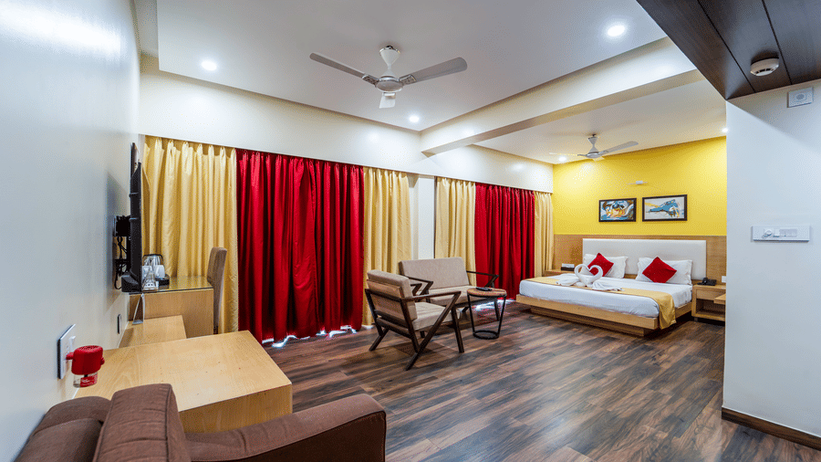 Rosetum Anjuna's Executive Suite with king bed, red and gold curtains, seating area, wall-mounted TV, wooden work desk, yellow accent wall, and dark hardwood floors.
