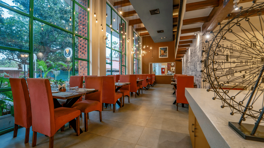 Rosetum Anjuna's ground floor restaurant with terracotta high-back chairs, wooden tables, Edison bulb pendant lights, floor-to-ceiling green-framed windows, and a mural wall.