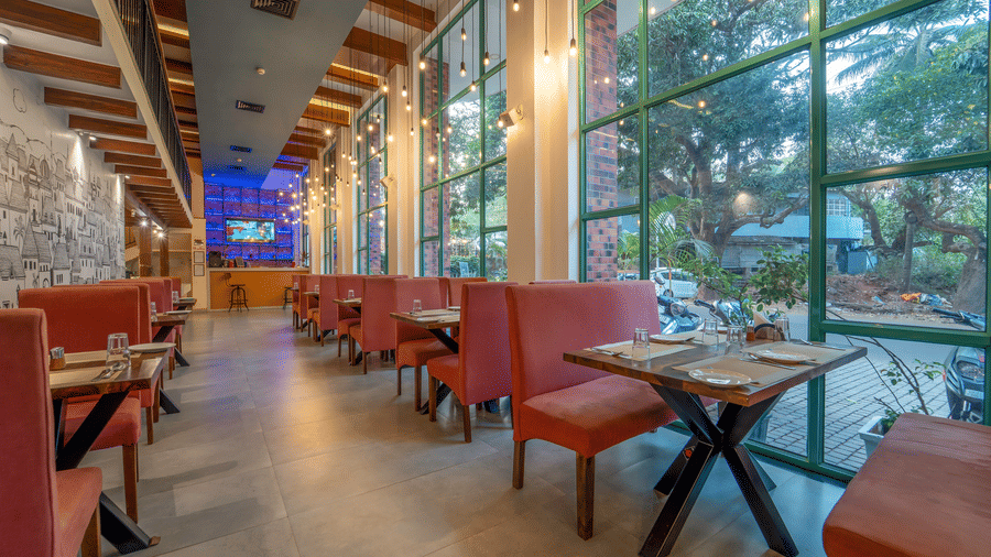 Rosetum Anjuna's restaurant dining area showing terracotta seating, pendant Edison lights, large glass windows with garden views, and an illustrated cityscape mural wall.